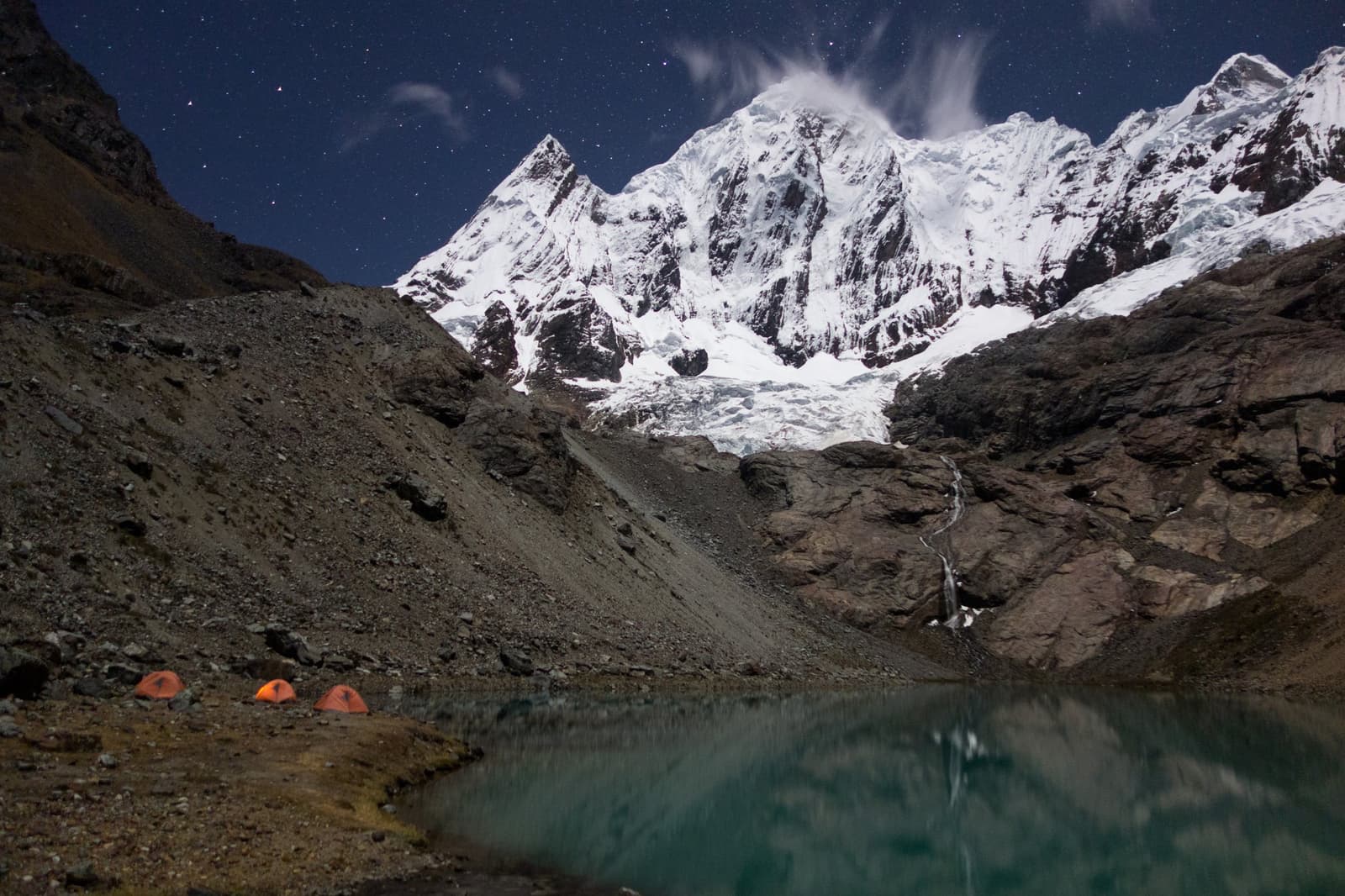 Stars out, glacier close, and those little tents look very small. Three orange tents pitched beside a glacial lake at night on the huayhuash circuit, with snow-covered peaks, a glacier tongue, and a star-filled sky reflected in the still water