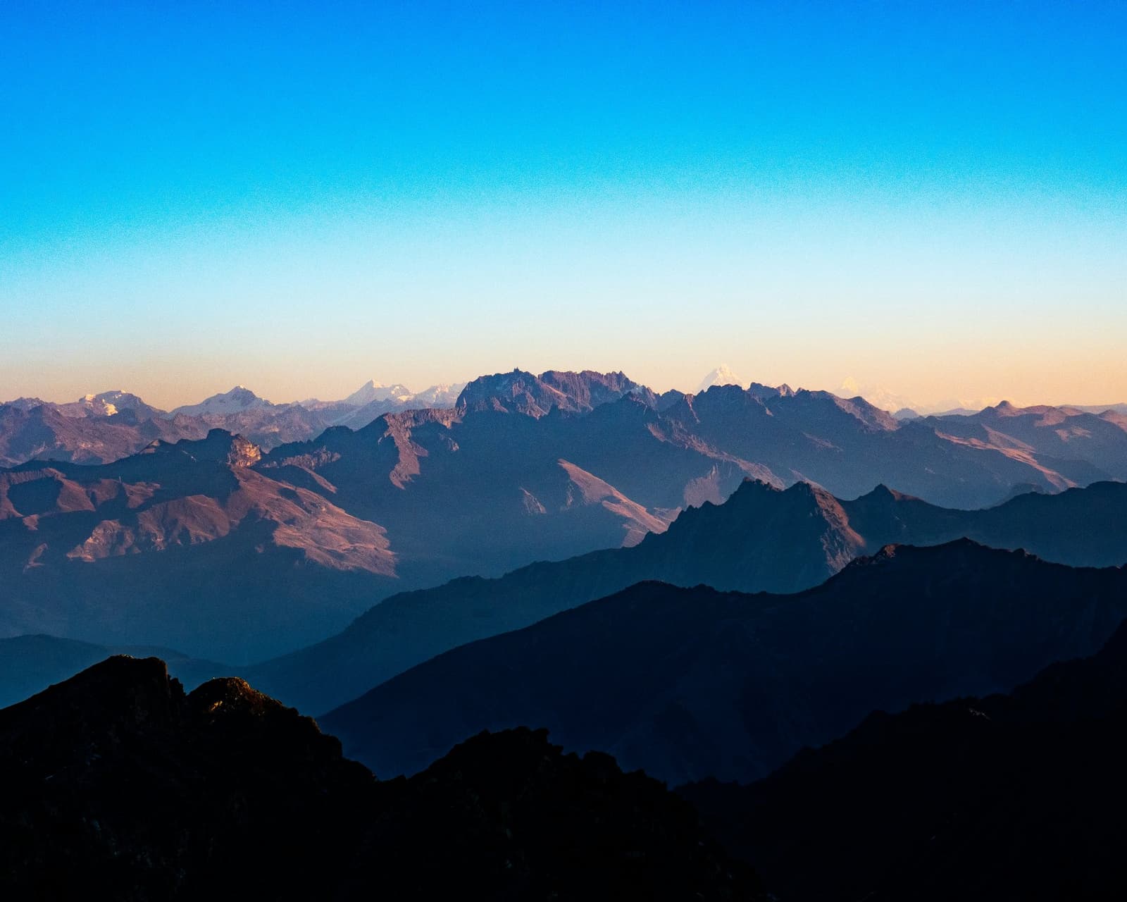 Ridge after ridge, and still more mountains behind those ones. Layered silhouettes of high Andean ridgelines recede into the distance under a clear blue sky fading to warm peach at the horizon, with snow-capped peaks visible on the cordillera huayhuash in the far background