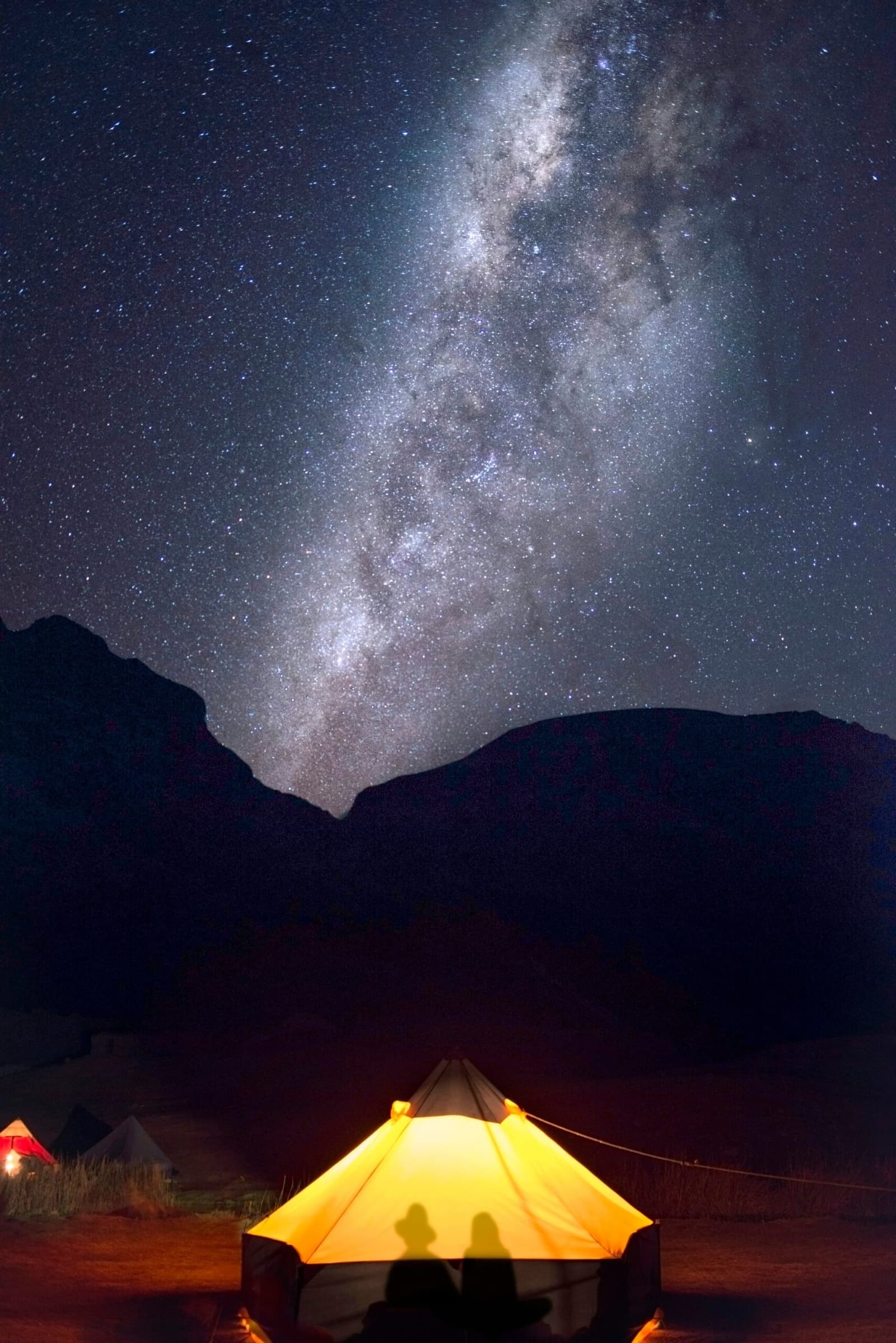 Your tent, a lantern. The galaxy, your ceiling. Sleep is optional. A glowing tent lit from within casts two silhouettes against the fabric, with the Milky Way arching over a dark mountain ridge on the huayhuash circuit