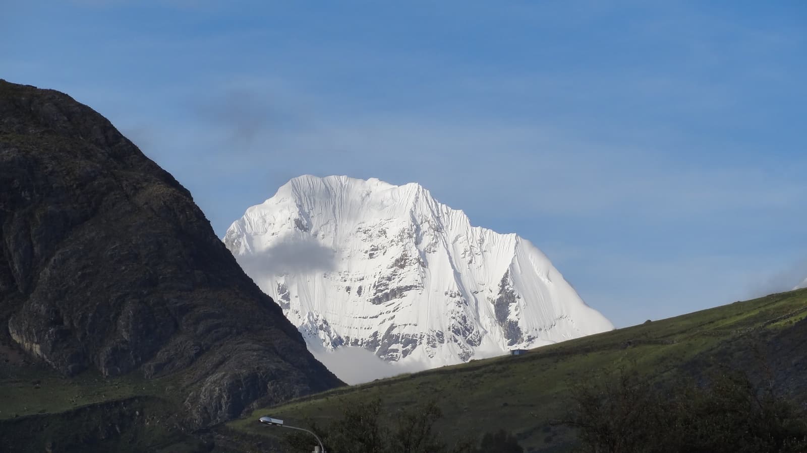 That much snow, that much silence, and the trail still goes higher. A broad, heavily glaciated snow-capped peak rises above green Andean hillsides on the huayhuash circuit, framed by a dark rocky ridge on the left under a clear blue sky