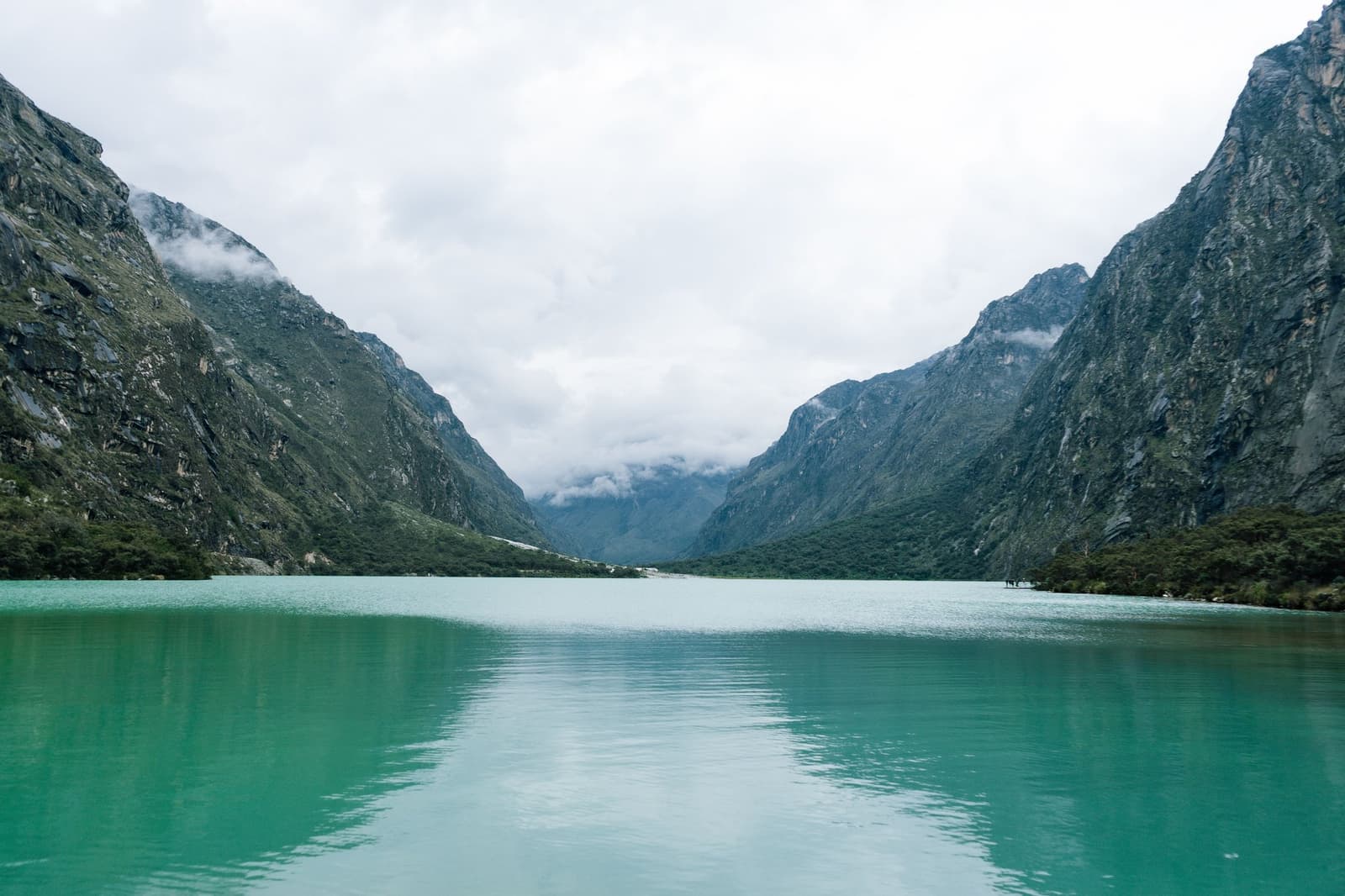 Esse verde pede uma segunda olhada antes de convencer. Vale a pena. Um lago glacial turquesa se estende entre duas encostas rochosas e íngremes sob um céu nublado no circuito Huayhuash, com nuvens baixas avançando pelo vale ao fundo