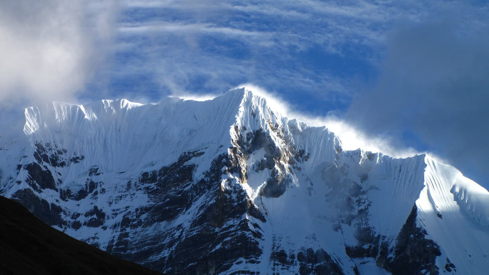 The ridge draws its own lines in the sky, and the wind does the rest. A snow-covered high-altitude peak on the huayhuash trek with wind-sculpted cirrus clouds swirling above the jagged glaciated ridgeline against a deep blue sky