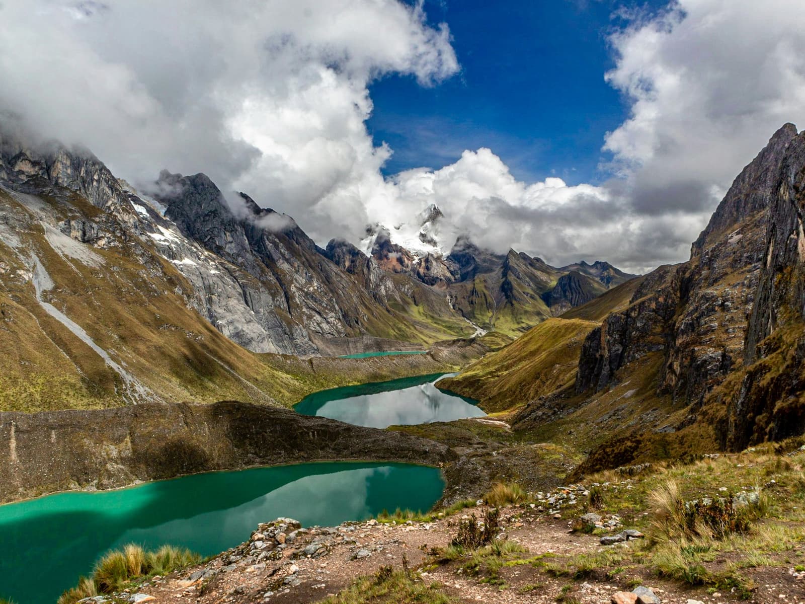 Three lakes, one valley, and a color you'll spend the rest of the trip trying to describe. A chain of turquoise glacial lakes fills a high Andean valley on the huayhuash trek, flanked by rugged rocky peaks and a snow-capped summit partially hidden by clouds under a blue sky