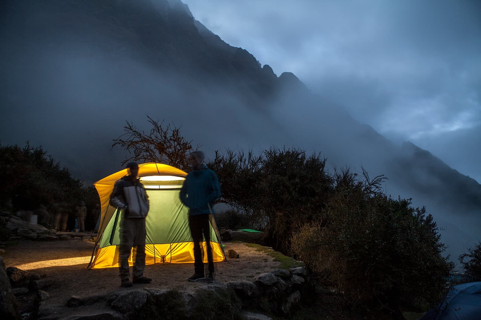 Cloud cover rolls in fast up here. Good thing the tent is already lit. Two trekkers stand outside a glowing yellow tent on the Inca Trail to Machu Picchu, with fog rolling over dark mountain peaks at dusk