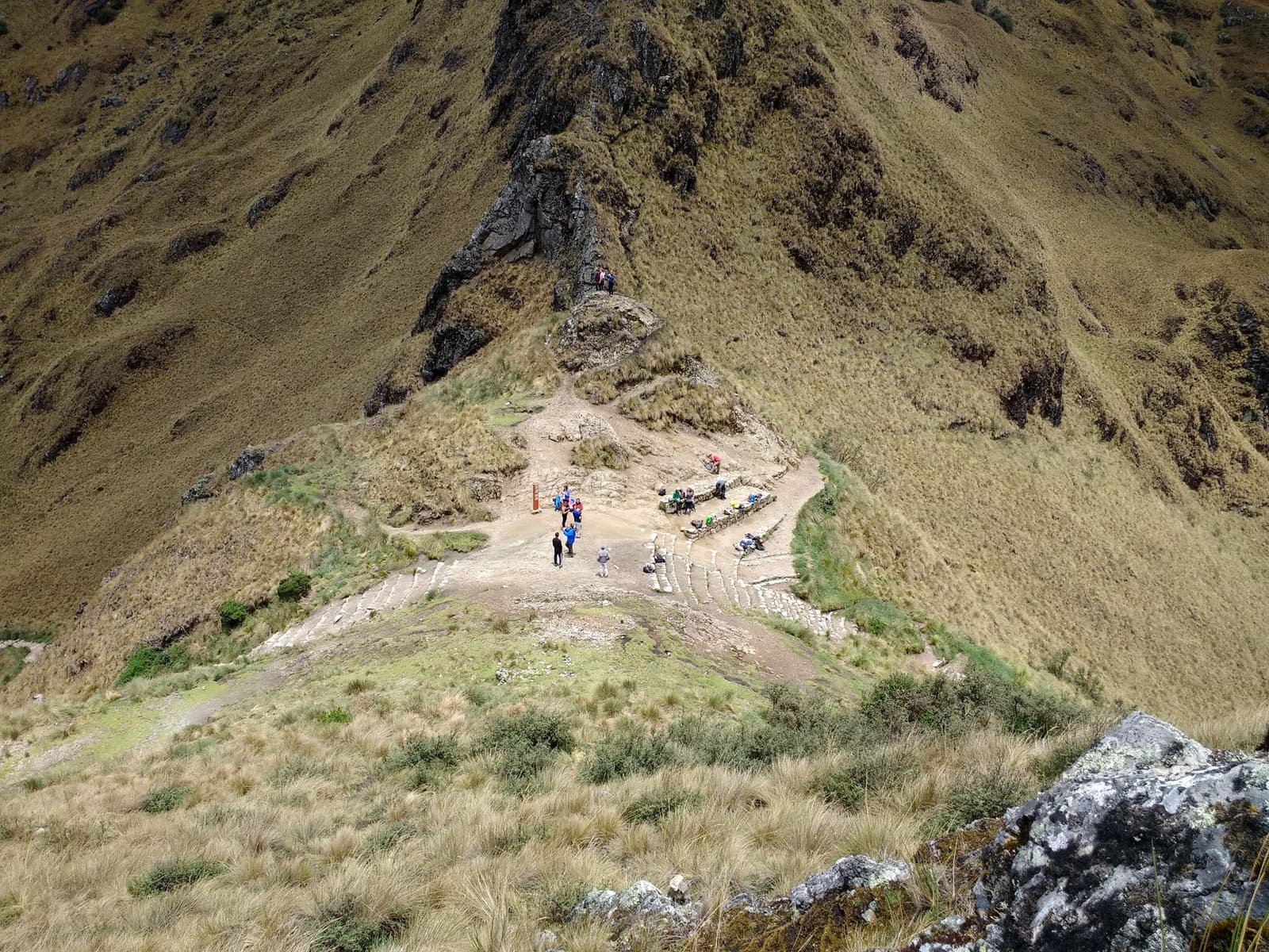 Somewhere between the last pass and the next one, legs vote for a break. Aerial view looking down at a rocky rest stop on the Inca Trail to Machu Picchu, with a group of trekkers pausing on stone-edged paths cut into a steep, grass-covered Andean hillside