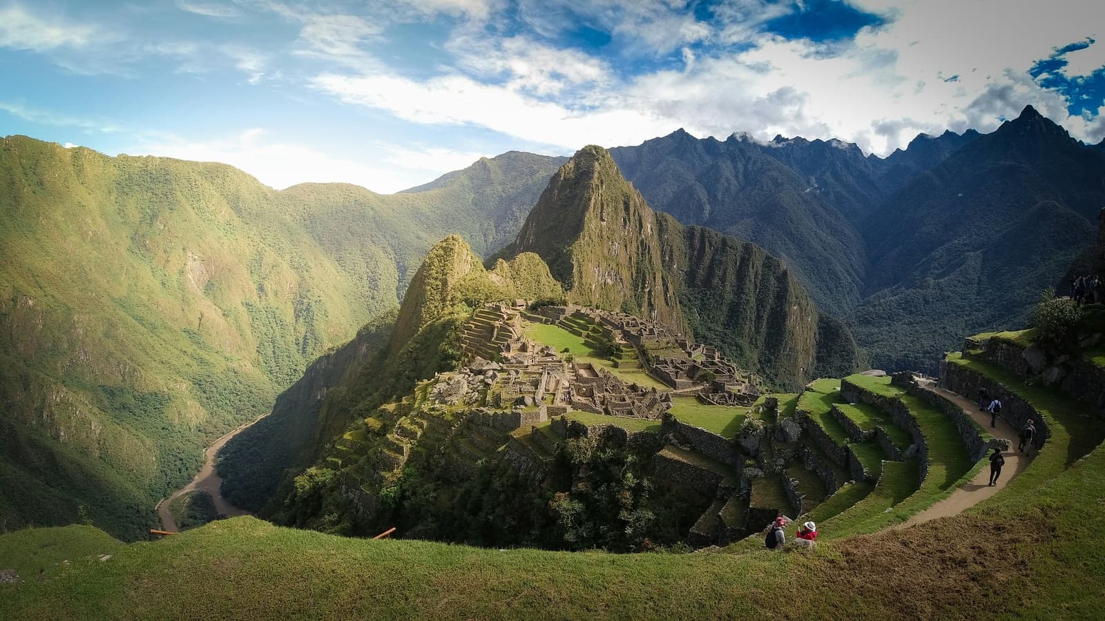 Six hundred years of stone, one ridge, and the whole valley spread out below you. Panoramic aerial view of Machu Picchu citadel on the inca trail to machu picchu, with Huayna Picchu peak rising behind the stone ruins, green agricultural terraces in the foreground, and the Urubamba River winding far below through the Andean mountains