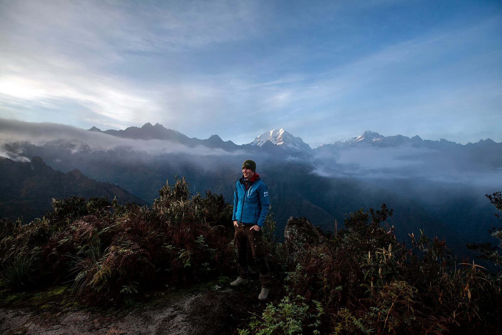 Cold air, big mountains, and the kind of smile that earns itself. A smiling hiker in a blue jacket and beanie stands on a high ridge along the Inca Trail to Machu Picchu, with cloud-draped Andean peaks and a snow-capped summit visible behind him