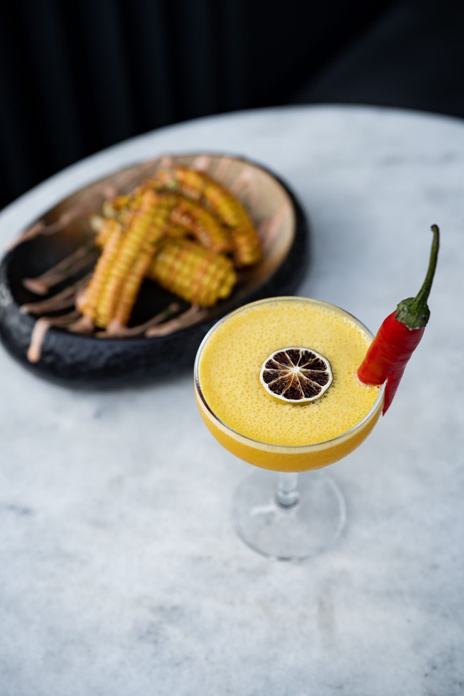 Spicy, citrusy, and nothing like trail mix. Lodge kitchens take their craft seriously. Photo: Kadir AVSAR A golden frothy cocktail in a coupe glass garnished with a dried lime wheel and a fresh red chili pepper, served alongside a dark bowl of grilled baby corn on a marble table, part of the gourmet dining experience on a luxury Salkantay trek