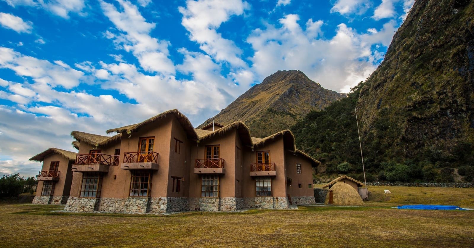 Your reward for the descent: a real bed, hot water, and that peak keeping watch outside. Colpa Lodge, a two-storey adobe and stone building with thatched roofs and wooden balconies, part of the luxury Salkantay trek lodge-to-lodge experience, set against a steep Andean peak and blue sky with scattered clouds