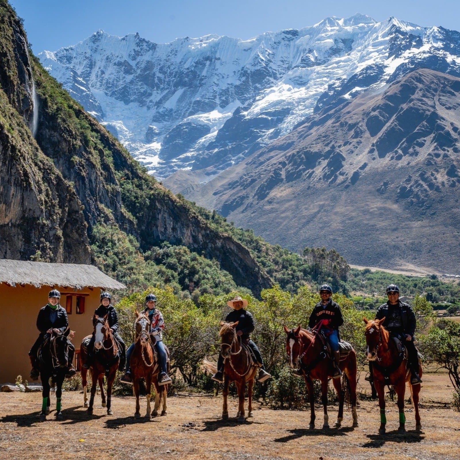 The optional horse ride hits different when the Andes put on a show like this. Six riders on chestnut horses pose for a group photo on the luxury Salkantay trek, with a snow-capped Andean peak, a cascading waterfall, and a traditional adobe building visible behind them