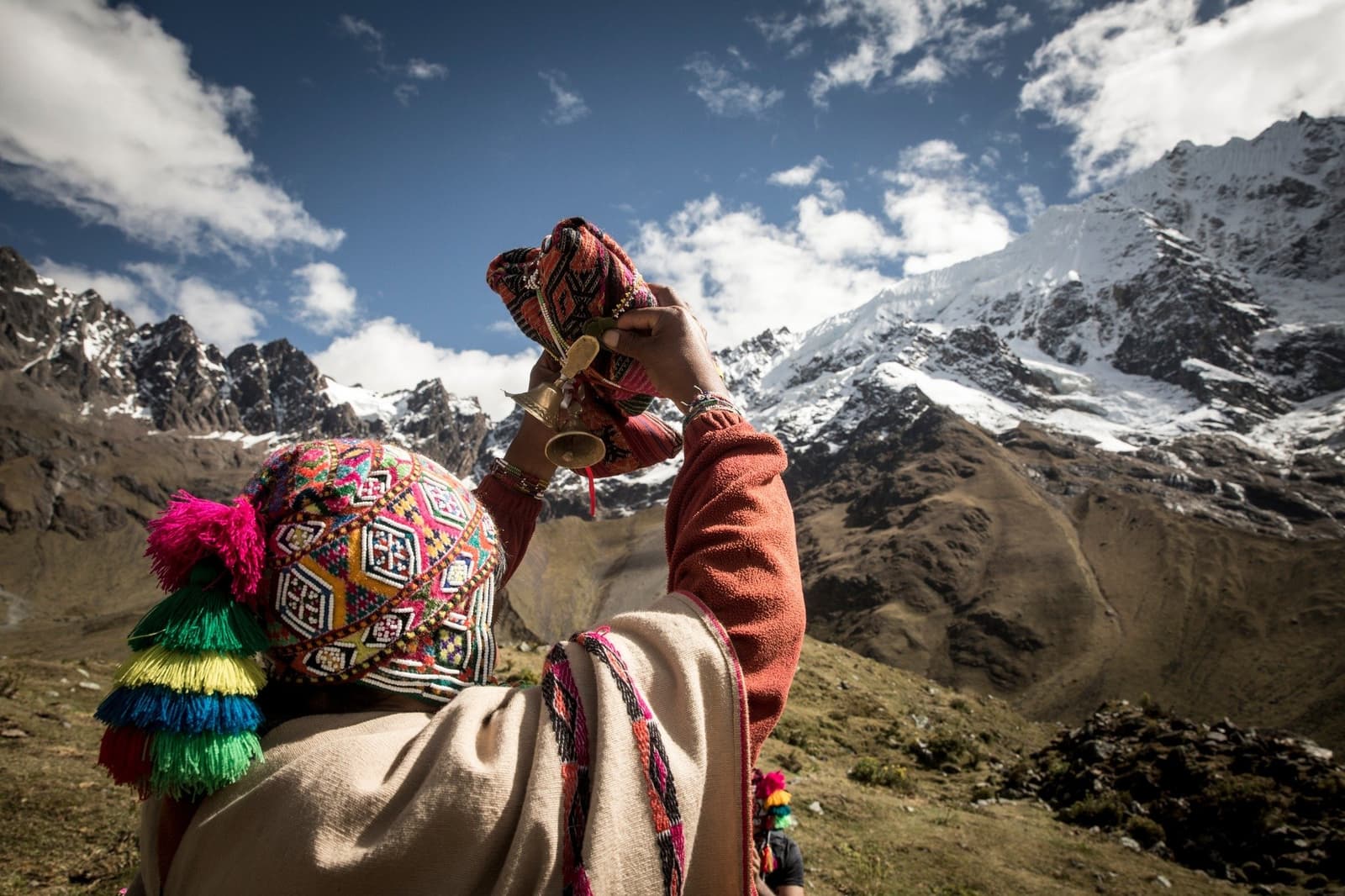 An offering to the mountain, small bell raised, glaciers listening. Person in a colorful traditional Andean woven hat and woven poncho holds a small bell and textile offering toward snow-capped Andean peaks during a Pachamama ceremony on this machu picchu adventure tour