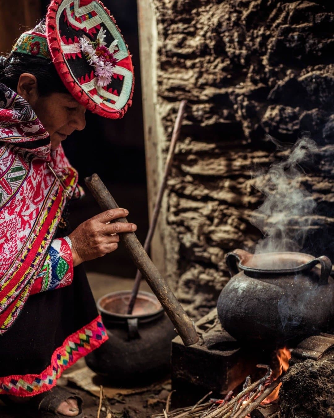 Lunch tastes different when it's cooked over a real fire, by someone who really knows how. A Quechua woman in traditional embroidered dress and flower-adorned hat stirs a steaming clay pot over an open wood fire during a machu picchu adventure tour community visit in the Andes