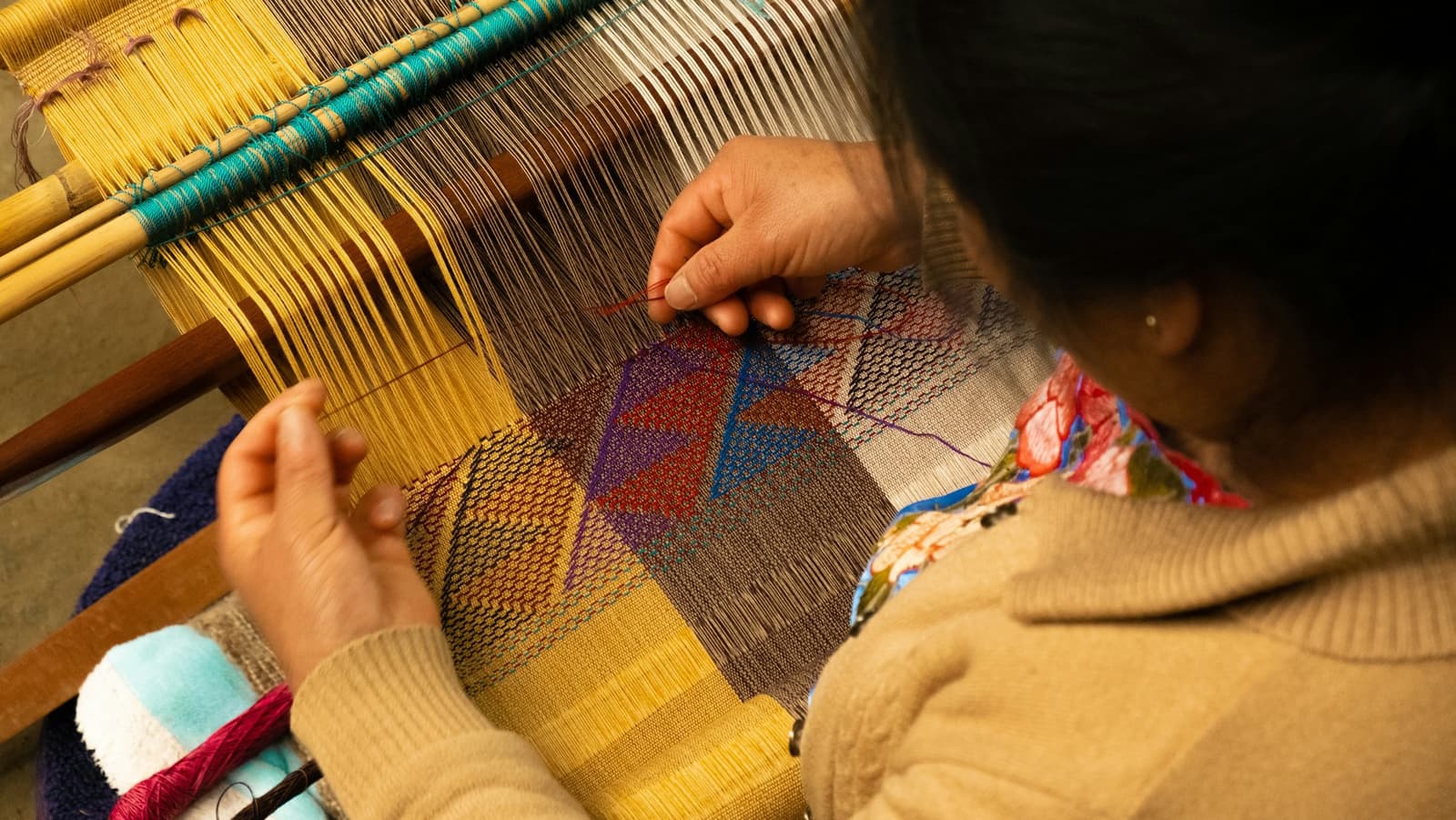 Each pass of the shuttle is a decision. The pattern doesn't write itself. Close-up overhead view of a woman's hands working a traditional wooden loom, weaving a multicoloured geometric pattern in yellow, red, purple, blue and grey threads, seen on a Machu Picchu and Amazon tour through Peru.