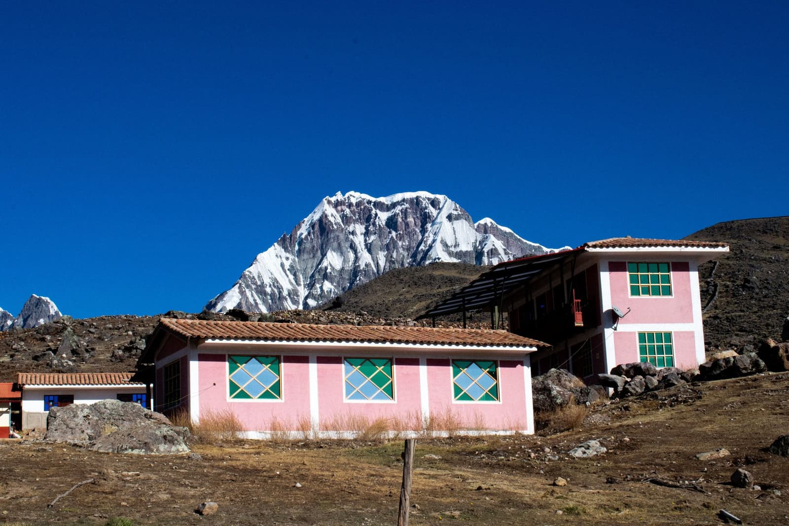 Pink walls, thin air, and a glacier that couldn't care less about altitude sickness. Pink-walled buildings with green-framed windows in a high-altitude Andean village, with a snow-capped rocky peak rising sharply against a deep blue sky on the overland crossing of this machu picchu amazon tour