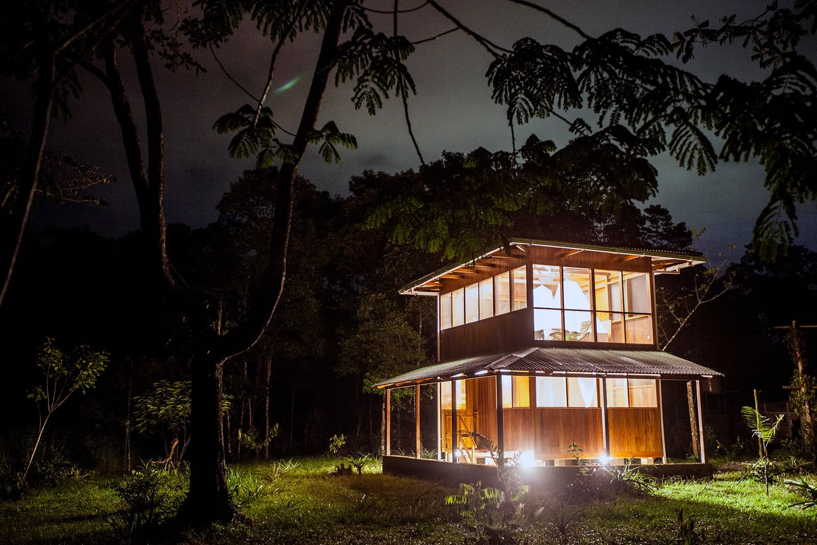 Nights in the cloud forest sound like a river and look like this. A two-storey wooden lodge glowing with warm interior light at night, surrounded by dense cloud forest trees on a Machu Picchu and Amazon tour through Peru