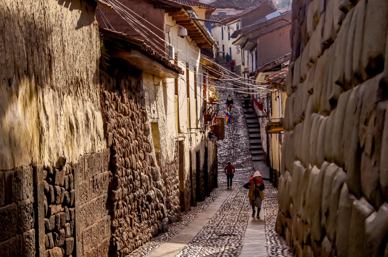 Cusco keeps two eras stacked on the same street, and somehow it works. Narrow cobblestone alley in Cusco flanked by ancient Inca stone walls, with pedestrians walking uphill past colonial buildings and colorful flags on this machu picchu amazon tour machu picchu and amazon tours route
