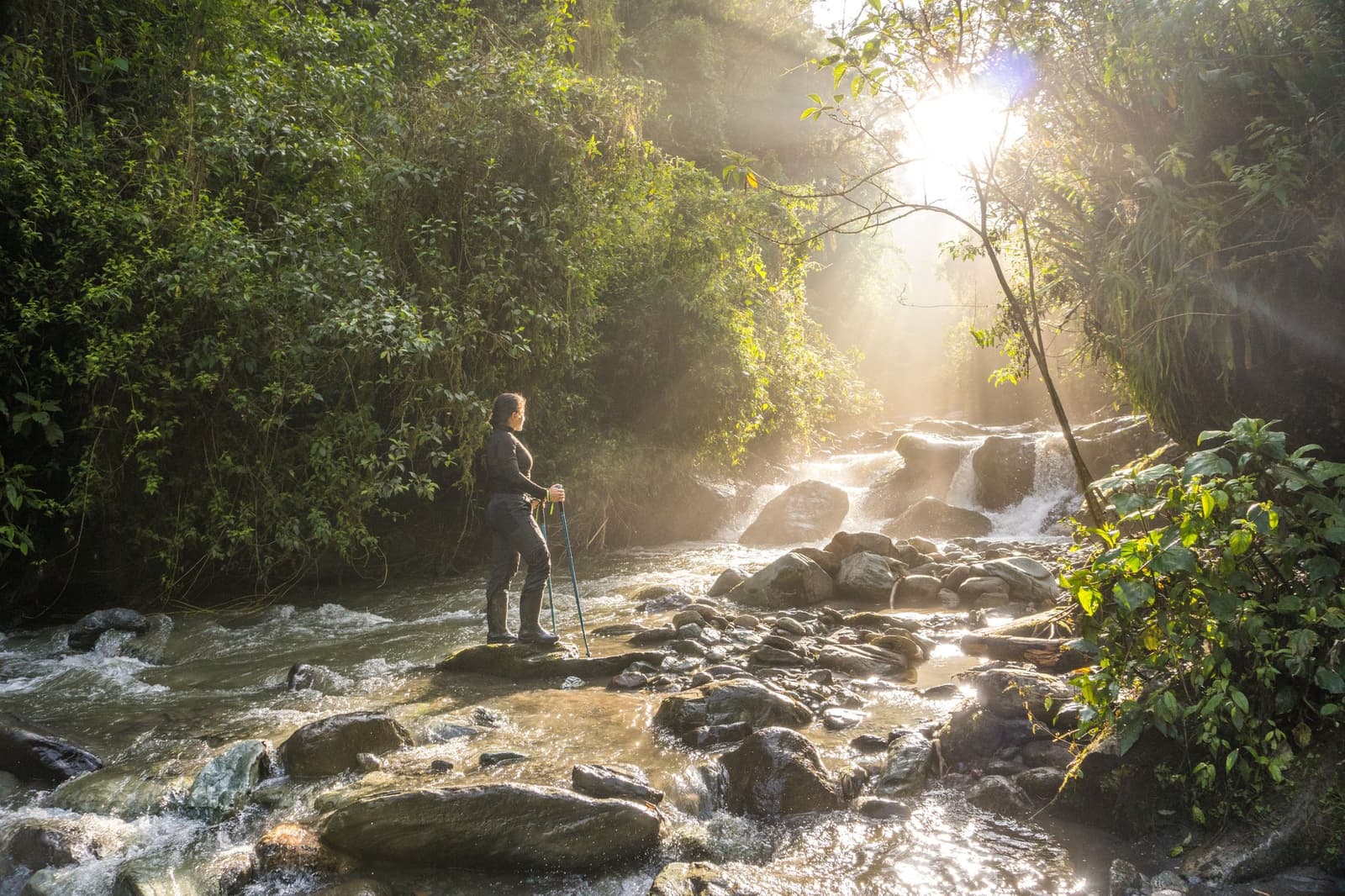 The Araza River doesn't pause for photos, but you will. A hiker with trekking poles stands on a boulder beside a rushing waterfall surrounded by dense green cloud forest, with sunlight streaming through the canopy on a Machu Picchu Amazon tour overland through Peru