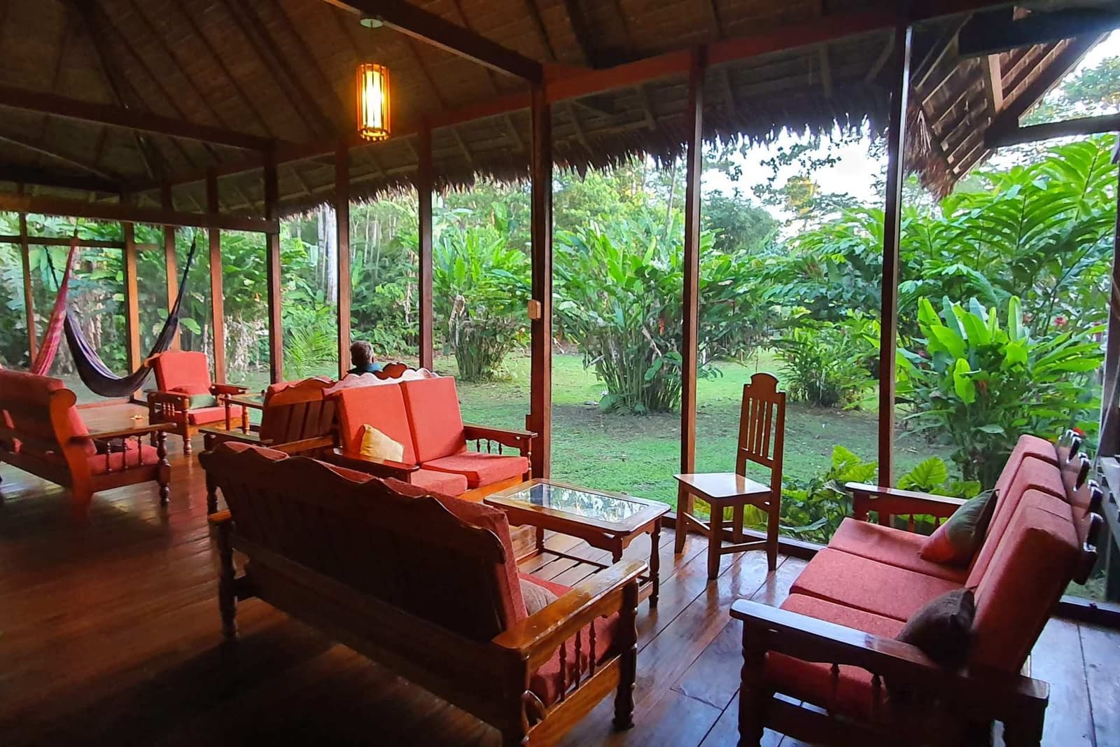 The kind of common room that makes you forget you had a plan for the afternoon. Open-sided jungle lodge lounge with red-cushioned wooden furniture, a hammock, thatched roof, and lush tropical garden views, part of a Machu Picchu and Amazon tour itinerary in the Peruvian rainforest