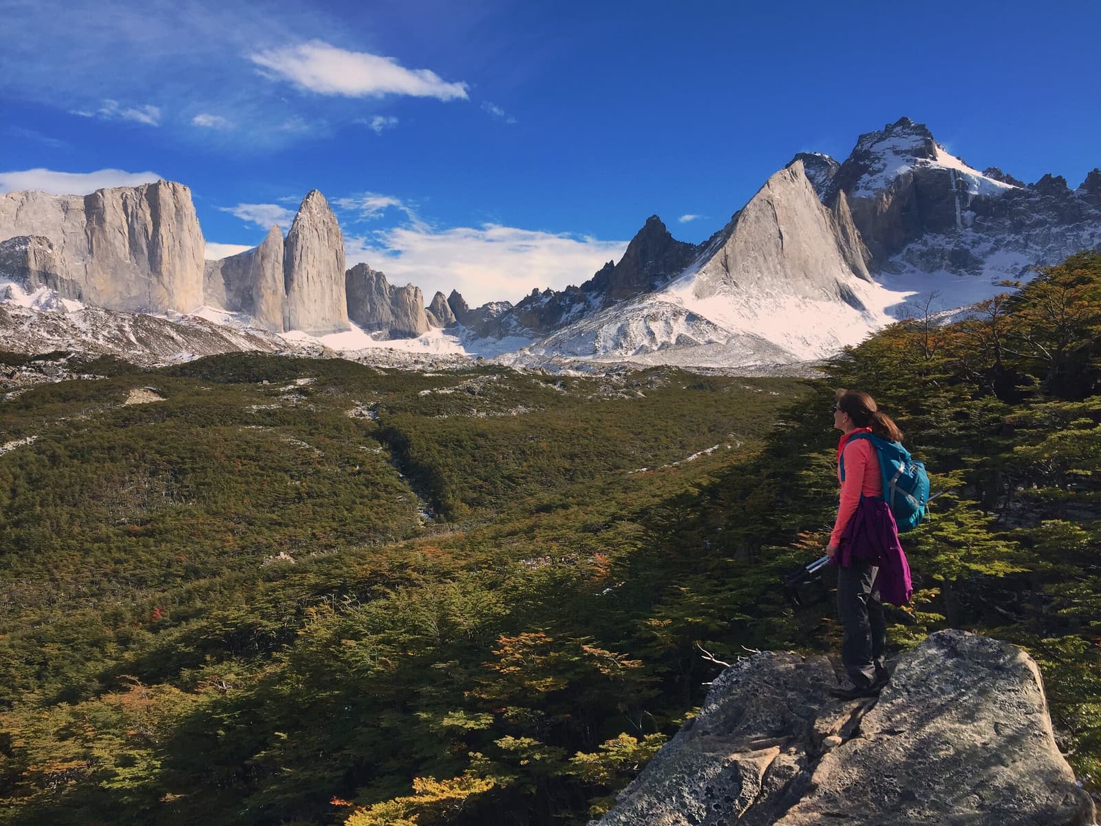 First day, first look at the Towers, and already the trip pays for itself. A female hiker with a blue backpack stands on a granite boulder, taking in the view of the three iconic Torres del Paine granite spires and snow-dusted peaks rising above a lenga beech valley on the self-guided O Circuit