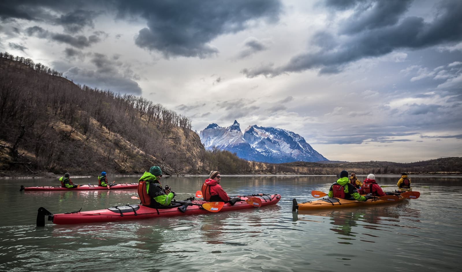 Some days the trail gives way to the water, and the mountains look even better from a paddle. A group of kayakers in red and orange sea kayaks paddle on a calm lake during the o circuit self guided trek, with the jagged snow-dusted peaks of the Cuernos del Paine rising sharply under a dramatic overcast sky