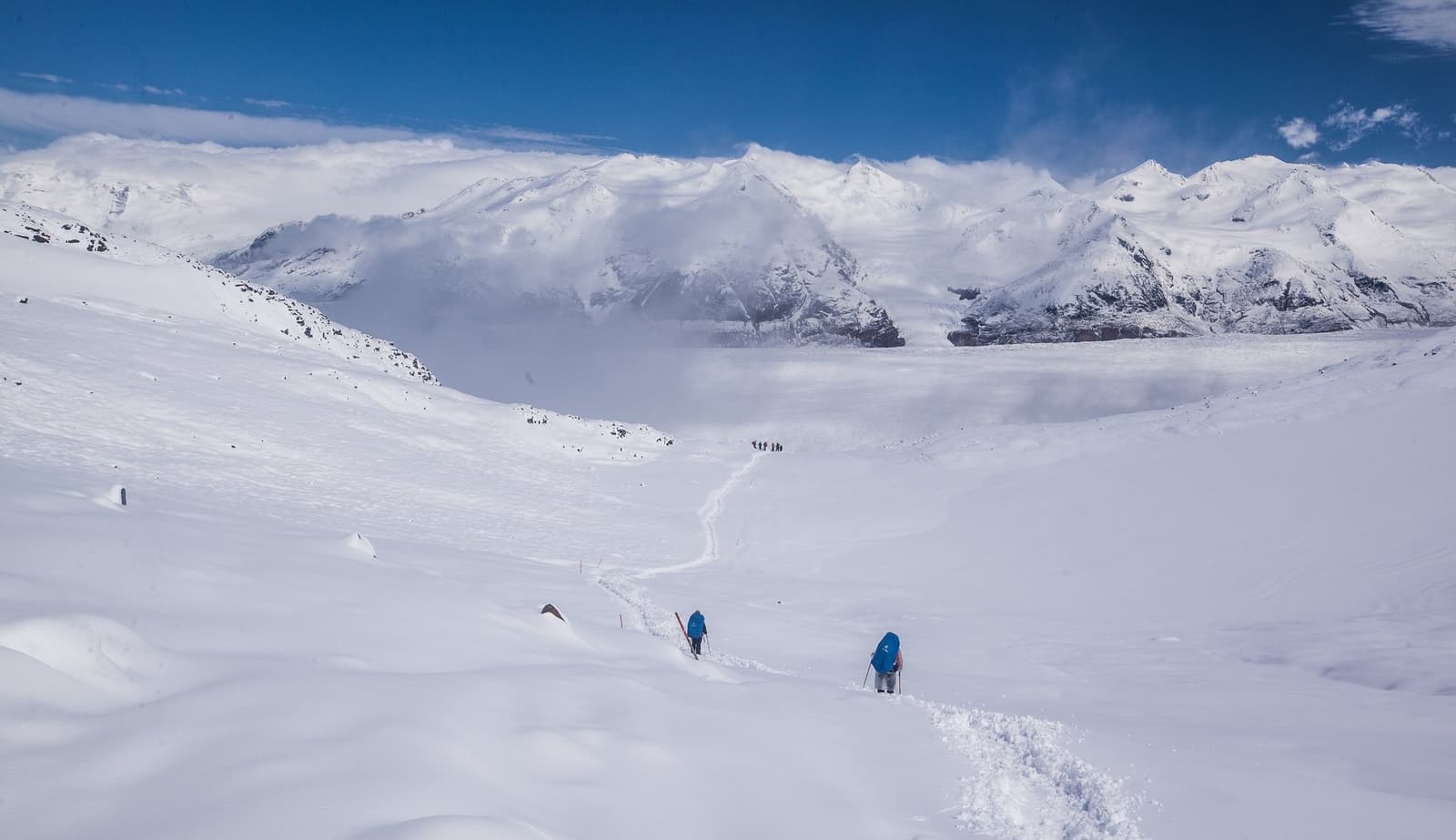 Two small figures, one big landscape. That ratio is exactly the point. Two trekkers with large blue backpacks follow a boot-packed trail across a vast snow-covered plateau on the self-guided O Circuit, with a sweeping range of snow-capped Patagonian peaks under a deep blue sky behind them