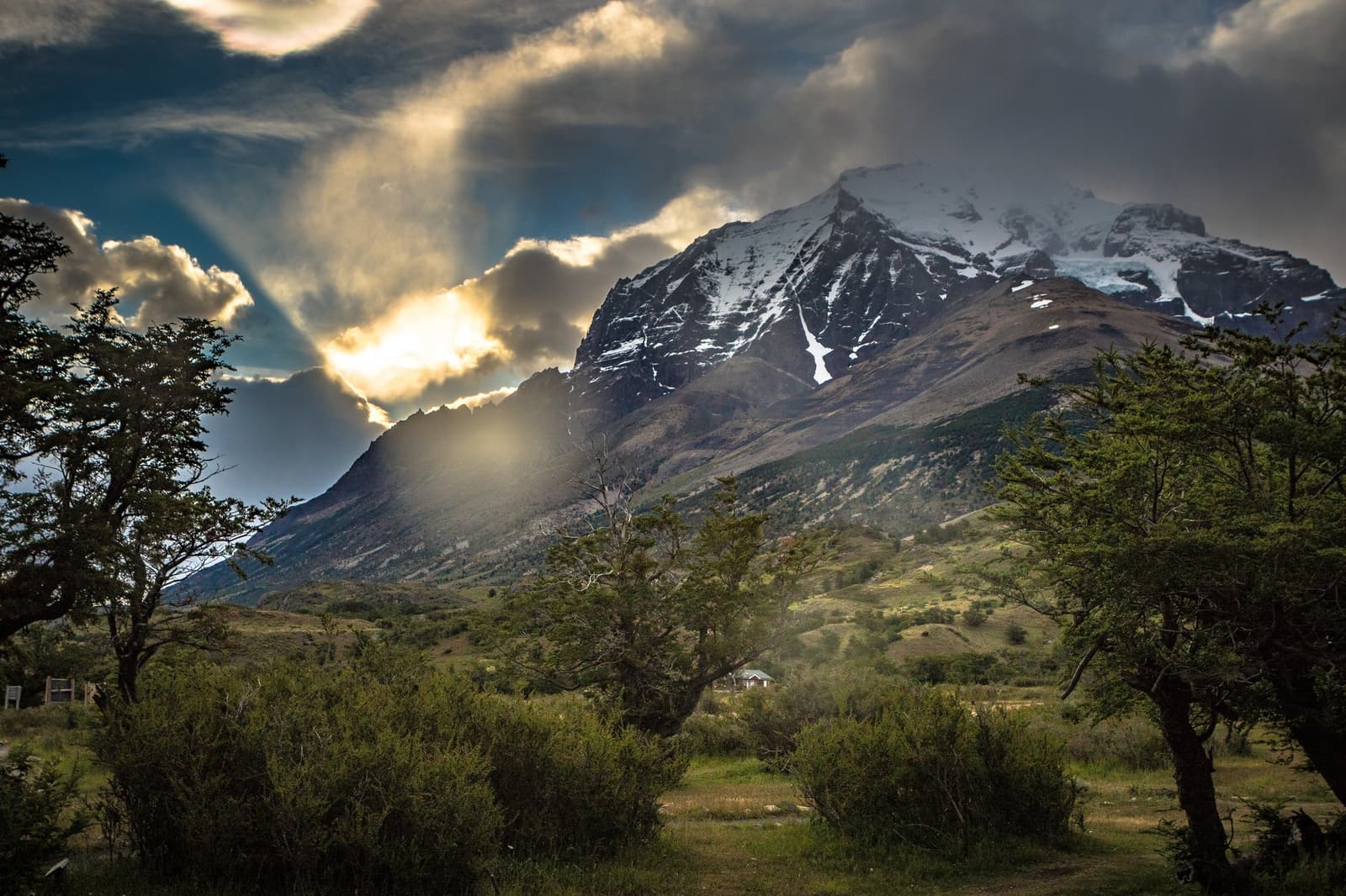 Sun through clouds, a small red shelter below, mountains doing the rest. Photo: outdoorstv.cl Snow-capped granite peak rising above a valley of lenga beech trees and green shrubland on the self-guided o circuit self-guided o circuit route in Torres del Paine, with sunlight breaking through dramatic storm clouds
