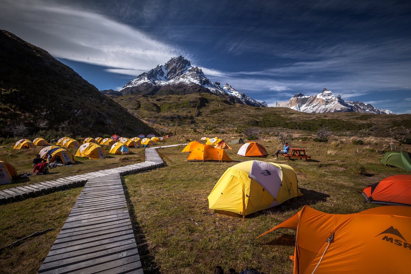 Your home for the night sorted, your next peak already in view. Dozens of yellow and orange tents spread across a grassy campsite on the self-guided O Circuit, with a wooden boardwalk path, snow-capped granite peaks, and the distinctive towers of Torres del Paine visible in the background under a deep blue sky.