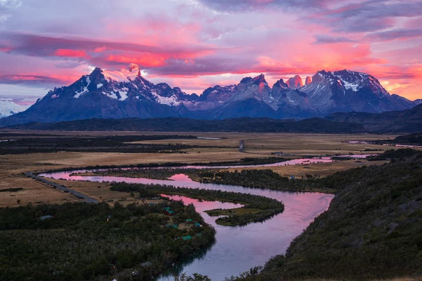 Wide aerial view of the self-guided O Circuit landscape at sunset, with a winding river reflecting pink and violet clouds, golden Patagonian steppe, and the snow-capped granite peaks of Torres del Paine glowing red in the last light Wide aerial view of the self-guided O Circuit landscape at sunset, with a winding river reflecting pink and violet clouds, golden Patagonian steppe, and the snow-capped granite peaks of Torres del Paine glowing red in the last light