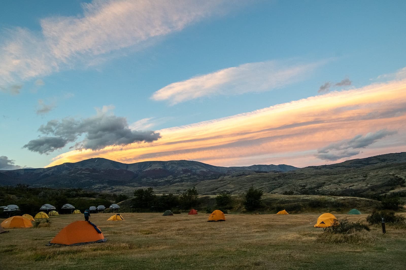 Good tents, good company, and that Patagonian sky doing whatever it wants. Colorful dome tents scattered across a dry grassland campsite on the o trek in patagonia, with warm pink and orange clouds streaking above a range of rounded mountains at dusk