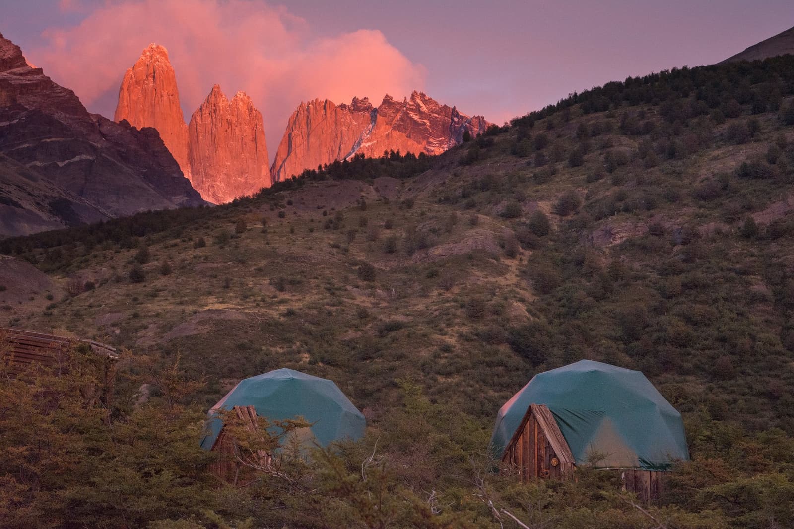 Your last two nights on the O Trek, and they happen to come with this view. Two geodesic dome shelters with wooden bases at EcoCamp Patagonia, with the granite towers of Torres del Paine glowing deep orange at sunset behind a forested hillside on the o trek patagonia circuit