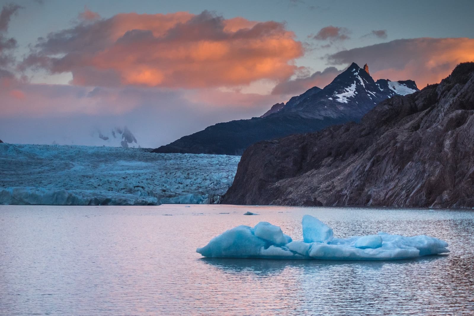 That iceberg calved recently. You can tell by the blue. A vivid blue iceberg floats on a calm glacial lake at sunset during the o trek in patagonia, with a large glacier face stretching to the left and snow-dusted rocky peaks glowing amber under orange-tinged clouds