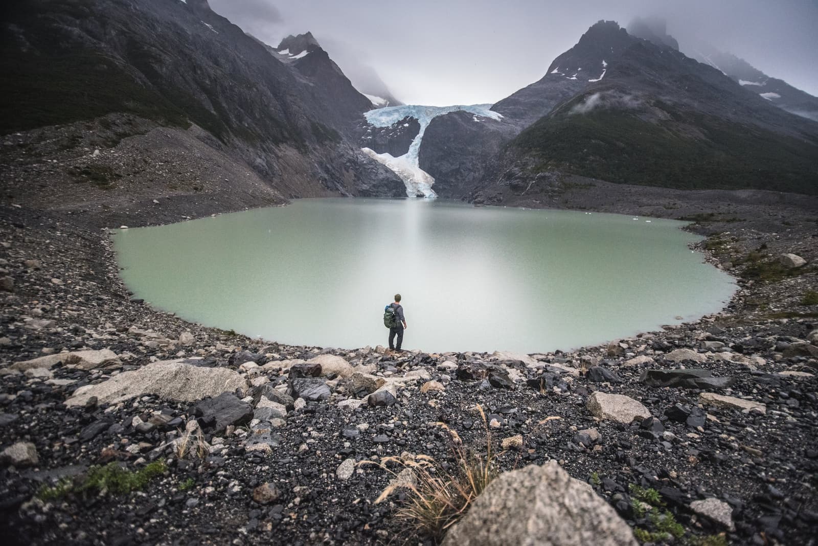 Still water, cold silence, and a glacier that has no interest in waiting. A lone hiker with a backpack stands at the rocky shore of a milky green glacial lake on the o trek in patagonia, facing a glacier that descends between dark, cloud-wrapped mountains