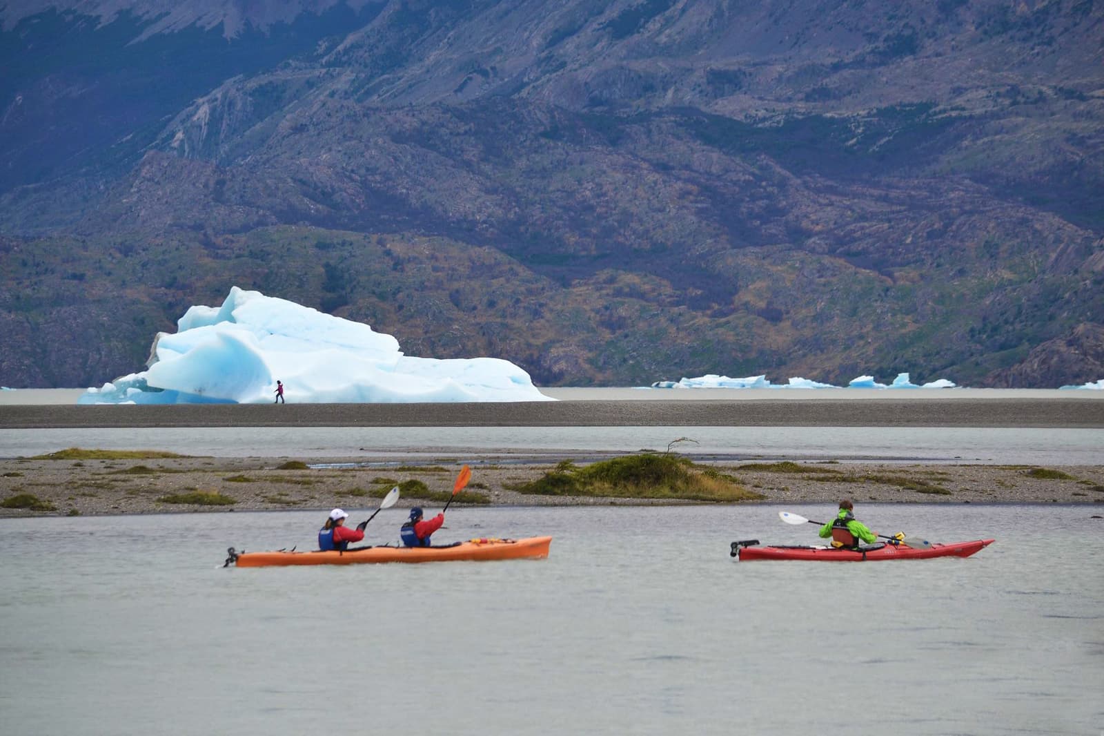 Paddles out, ice at eye level, mountains doing the rest. Three kayakers in orange and red kayaks paddle across a glacial lake on the o trek in patagonia, with a large blue iceberg and rugged mountain slopes visible behind them