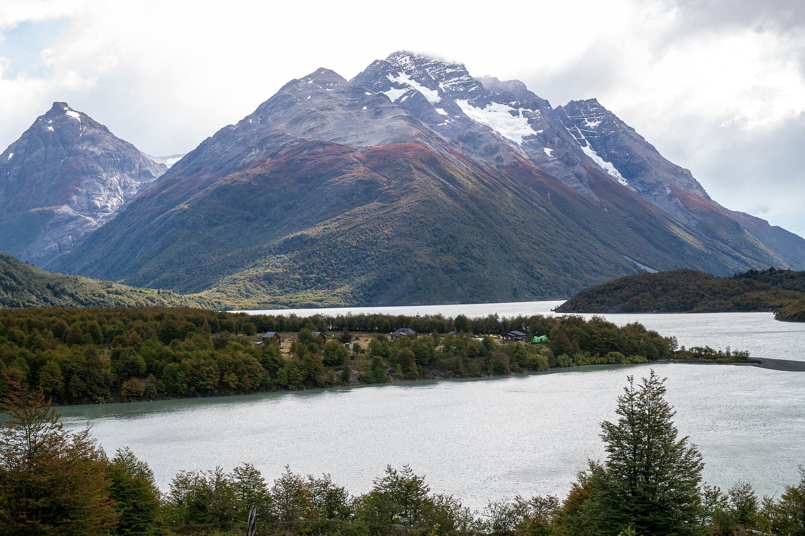 A campsite this remote earns its views fair and square. A calm glacial lake reflects grey skies in front of a massive snow-capped peak streaked with rust-red and green lenga forest, with a small cluster of wooden lodge buildings nestled on a forested peninsula — a scene from the o trek in patagonia's remote northern circuit.