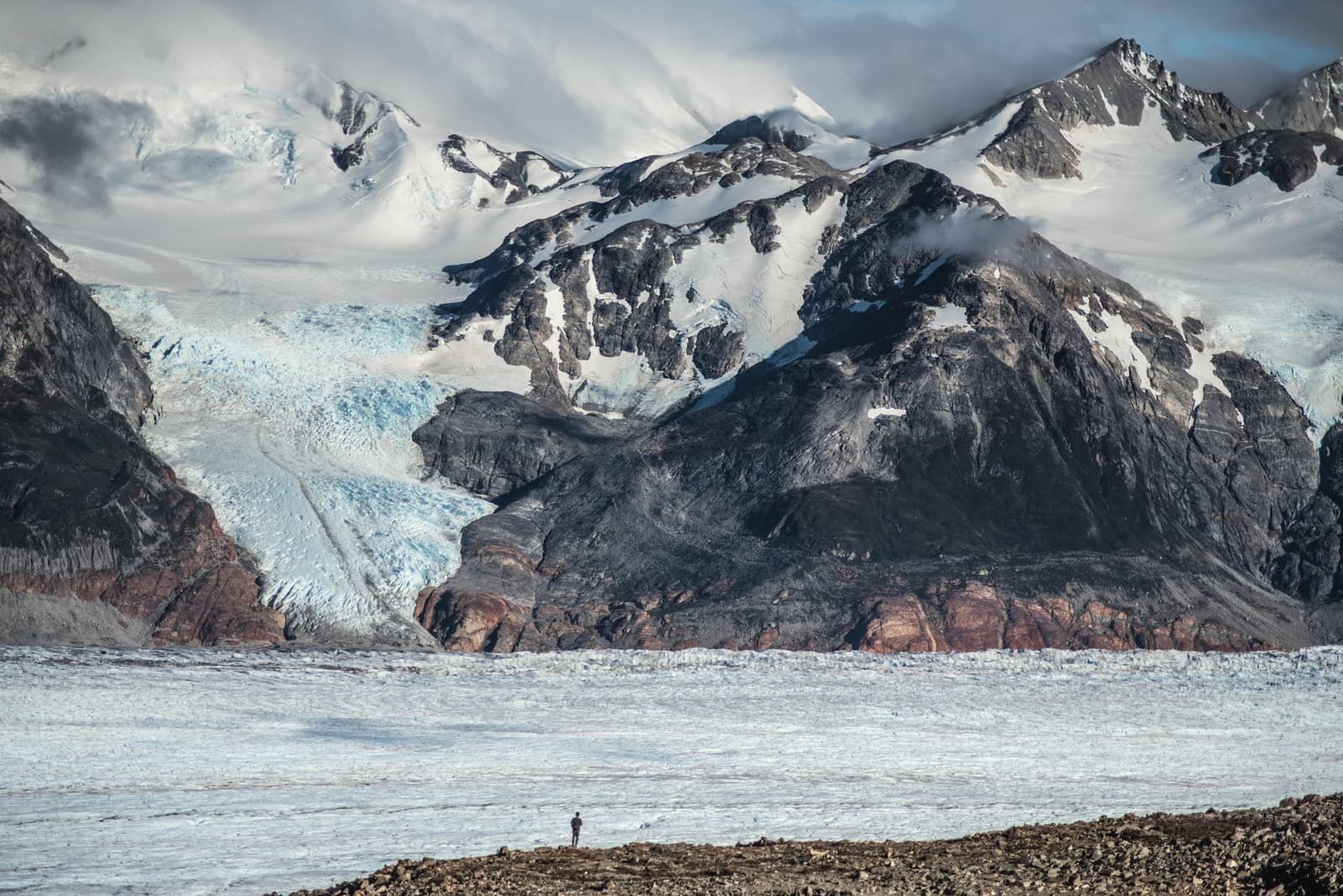 One person, one glacier, and the full weight of how far you've come. A lone hiker stands on a rocky moraine in front of a vast glacial ice field on the O trek in Patagonia, with a blue glacier tongue and snow-dusted peaks rising behind