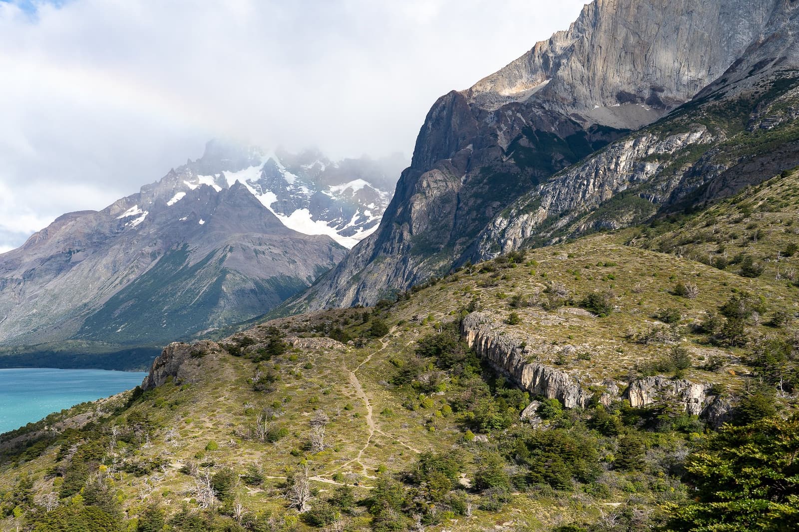That thin line cutting through the scrub is your path, and yes, it goes all the way up there. A narrow dirt trail winds through low Patagonian scrubland along the o trek patagonia route, with a turquoise glacial lake visible to the left and massive snow-dusted granite peaks rising into cloud above