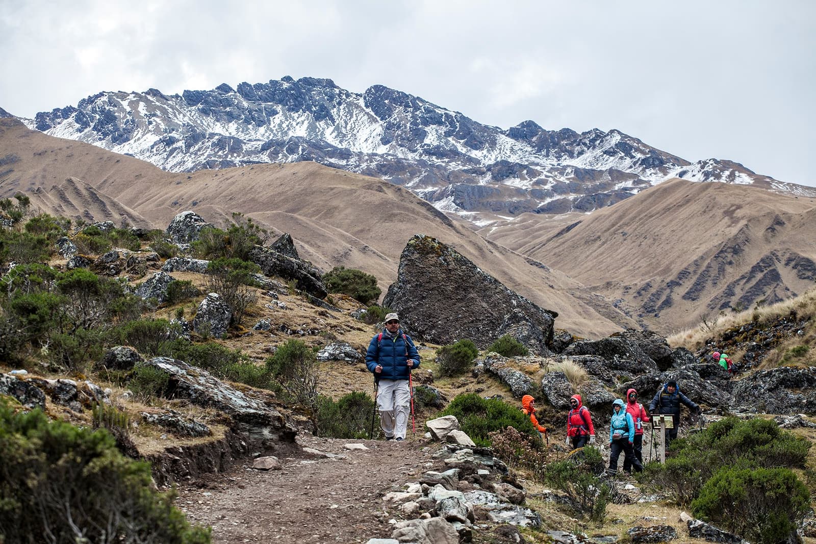 Layers of altitude, one trail, and a whole lot of puffer jackets. A group of trekkers on the off-the-beaten-path Salkantay trek hiking a rocky high-altitude trail, with snow-dusted peaks and sweeping brown ridgelines filling the background under an overcast sky