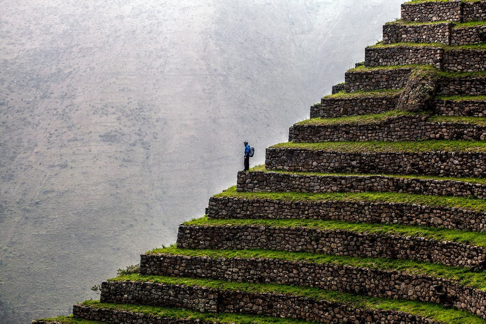 Small figure, enormous walls, the Incas built to last, and the mist knows it. A lone backpacker in a blue jacket stands on the green grass-covered stone agricultural terraces along the off-the-beaten-path Salkantay trek, dwarfed by the massive stacked retaining walls rising into misty mountain air