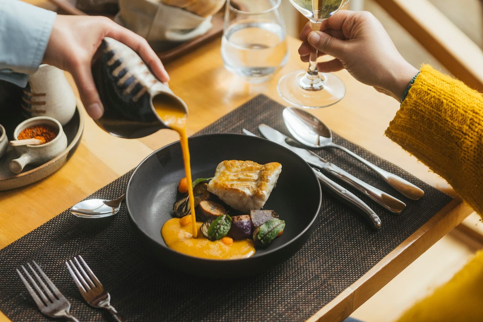 Sauce arriving tableside, wine already in hand. Dinner has its priorities straight. Photo: Patagonia Camp A server pours a vivid golden sauce from a ceramic pitcher over a pan-seared white fish fillet with roasted vegetables in a matte black bowl at Patagonia Camp, while a guest holds a glass of white wine nearby