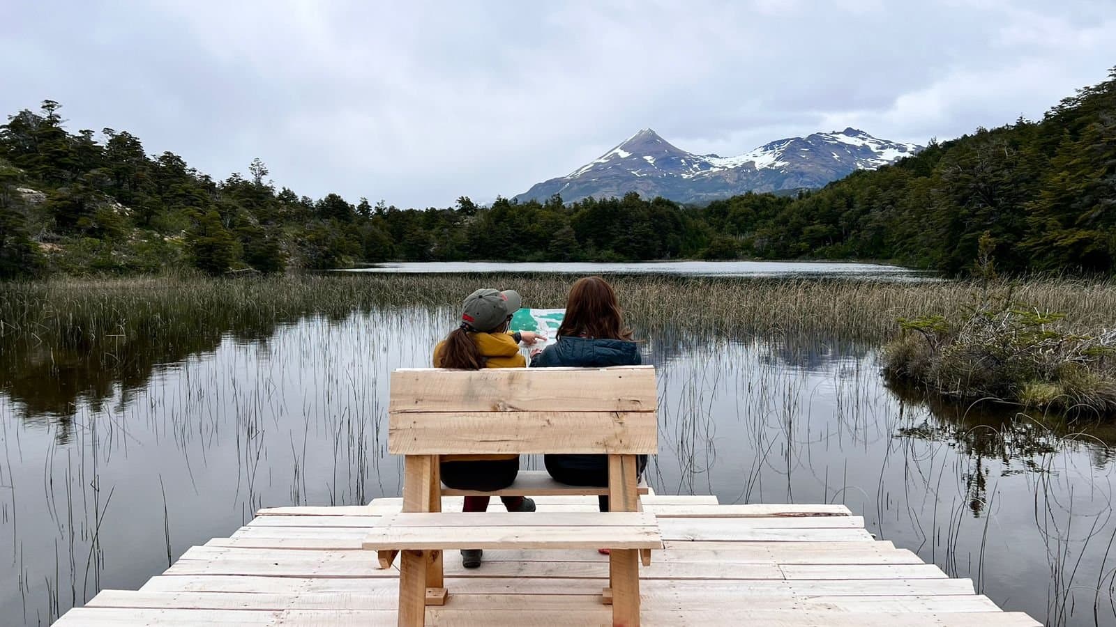 Planning the next few days with a view doing most of the convincing. Photo: Patagonia Camp Two travellers sit side by side on a wooden bench on a lakeside dock at Patagonia Camp, reading a map together, with a calm reed-fringed lake, lenga forest, and a snow-capped pointed peak reflected in the still water behind them