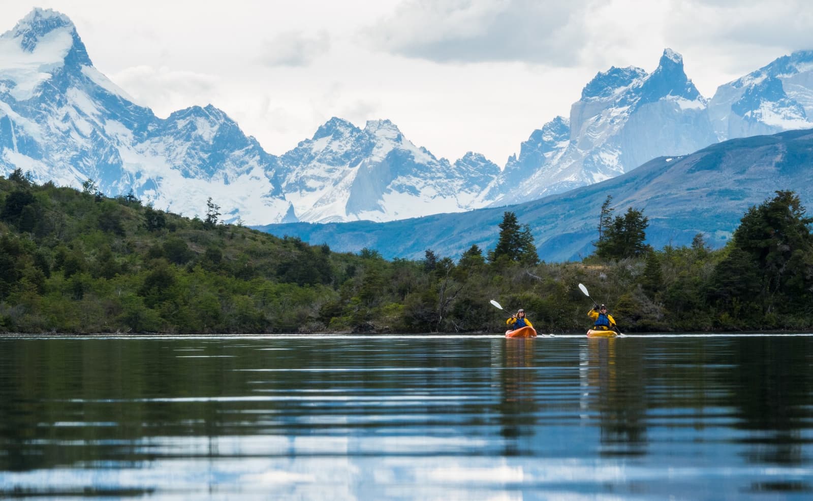 Paddles out, mountains ahead, and not a single queue in sight. Photo: Patagonia Camp Two kayakers in orange and yellow kayaks paddling on Lake Toro during a Patagonia Camp excursion, with the snow-capped peaks of the Paine Massif rising behind a fringe of native forest