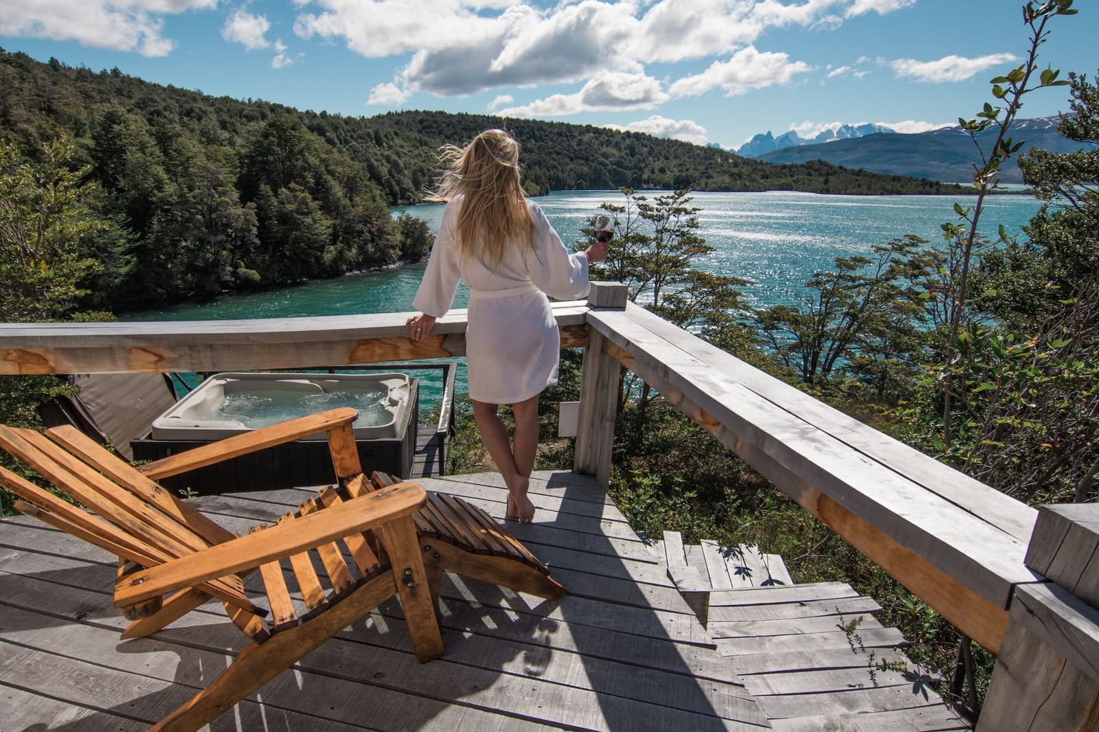 Wine in hand, robe on, wind doing whatever it wants, some afternoons need no agenda. Photo: Patagonia Camp A guest in a white bathrobe holds a glass of red wine on a private wooden deck at Patagonia Camp, with a hot tub, Adirondack chairs, and a turquoise glacial lake framed by forested hills and distant snow-capped peaks behind her