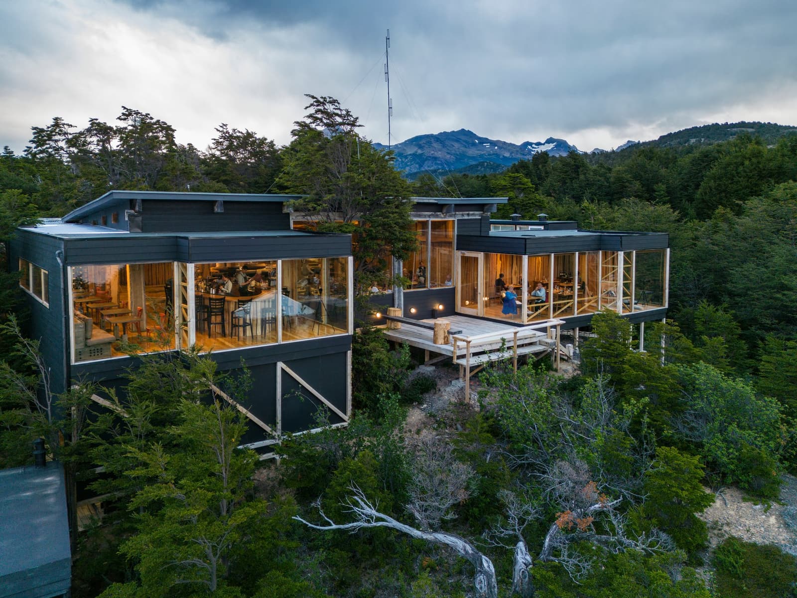 Dinner is on, the windows are lit, and the forest has no idea how good the wine list is. Photo: Patagonia Camp Aerial view of the Patagonia Camp lodge at dusk, two dark-clad glass-walled pavilions glowing with warm interior light, set among native lenga beech trees on a forested hillside, with snow-dusted mountains rising under a cloudy sky in the background.