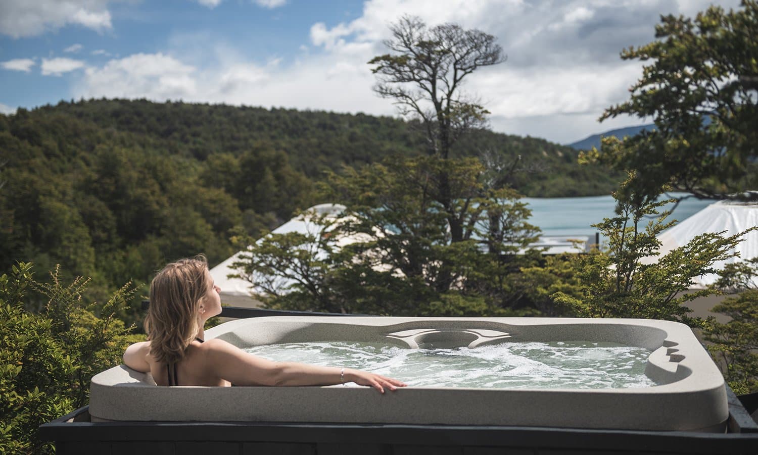 Hot water, cold air, and a lake that has no interest in being rushed. Photo: Patagonia Camp A guest relaxes in an outdoor jacuzzi at Patagonia Camp, gazing through native lenga forest toward the turquoise waters of a lake and forested hills under a partly cloudy sky