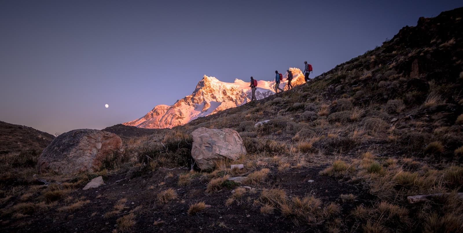First light on the snow, and your boots are already moving. A good way to start a day. Photo: Patagonia Camp Three hikers with backpacks ascend a rocky hillside at dawn on a patagonia camp excursion, with a snow-dusted mountain peak glowing amber in alpenglow and a full moon hanging in the blue-violet sky above
