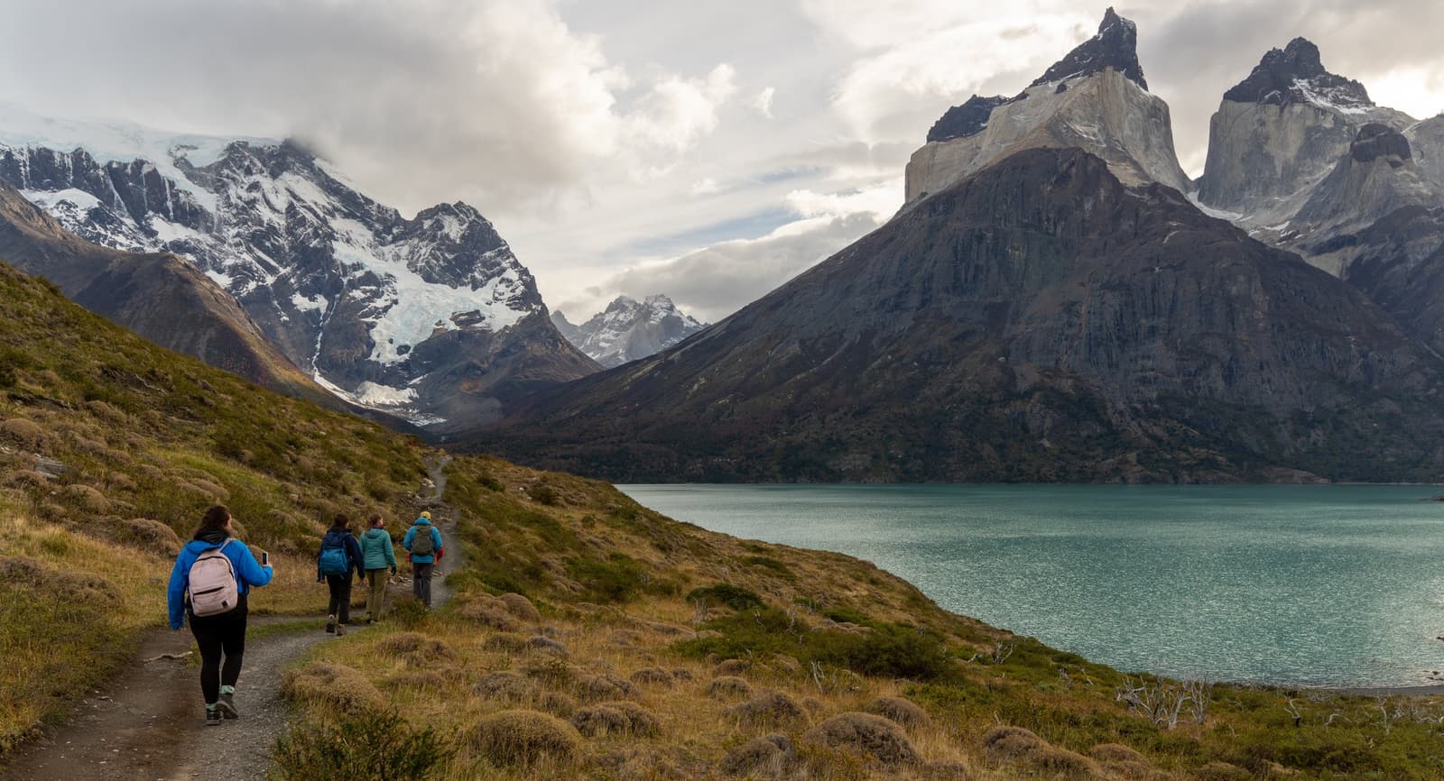 The path keeps going, and so do you, the Massif has a way of pulling you forward. Photo: Patagonia Camp Four hikers with backpacks walk a dirt trail along the shore of a glacial lake on a patagonia camp excursion, with the granite towers and snow-capped peaks of the Paine Massif rising dramatically in the background under a cloudy sky