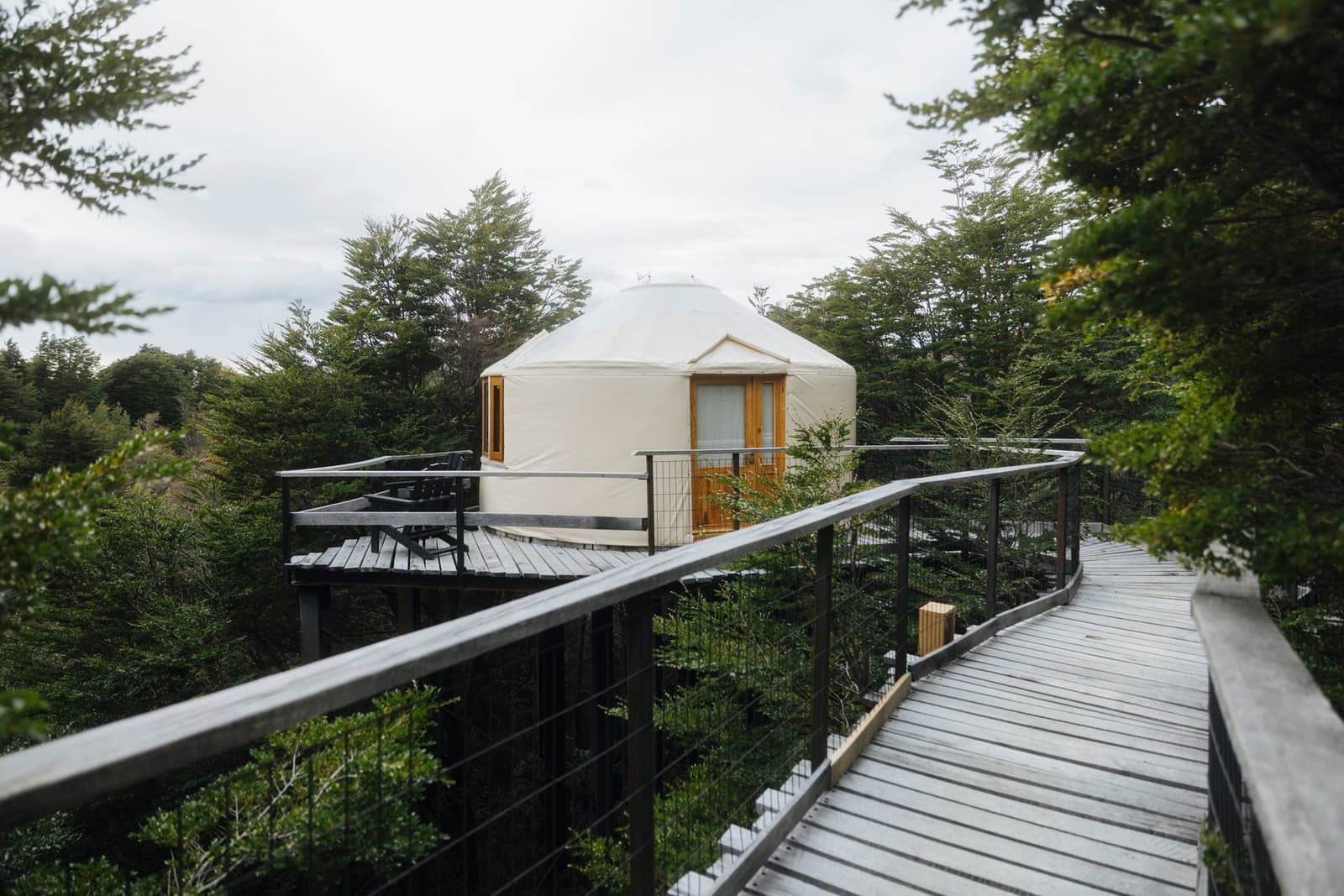 Your room is up in the trees, and the boardwalk makes the journey half the fun. Photo: Patagonia Camp A white circular yurt at Patagonia Camp sits on a raised wooden deck above a native forest canopy, reached by a curving elevated boardwalk with cable railings