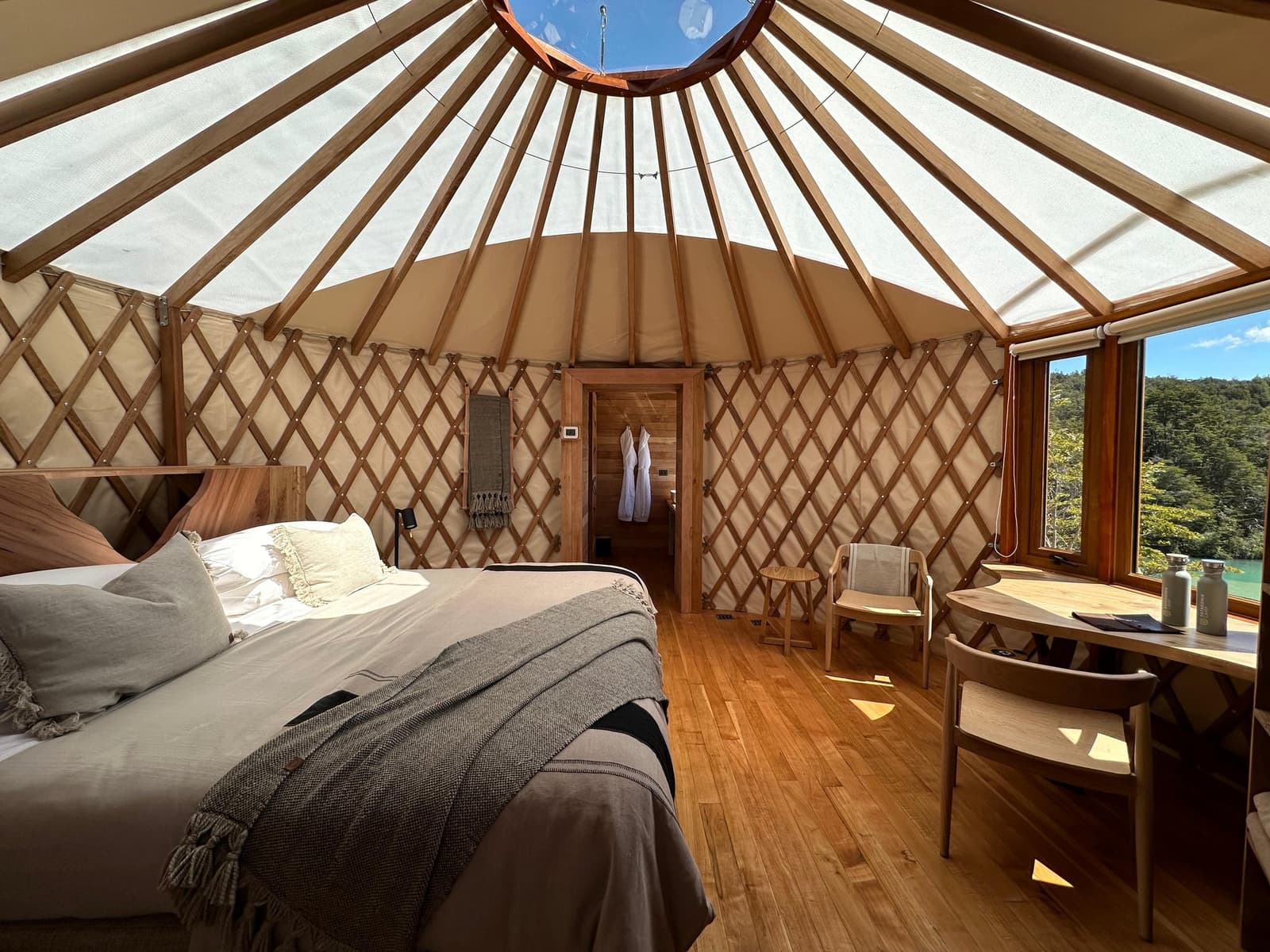 More headroom than most apartments, and a skylight that doubles as an alarm clock. Photo: Patagonia Camp Interior of a patagonia camp yurt with a king bed, warm wood lattice walls, a translucent domed roof with a central skylight, and a window framing green forest and blue sky