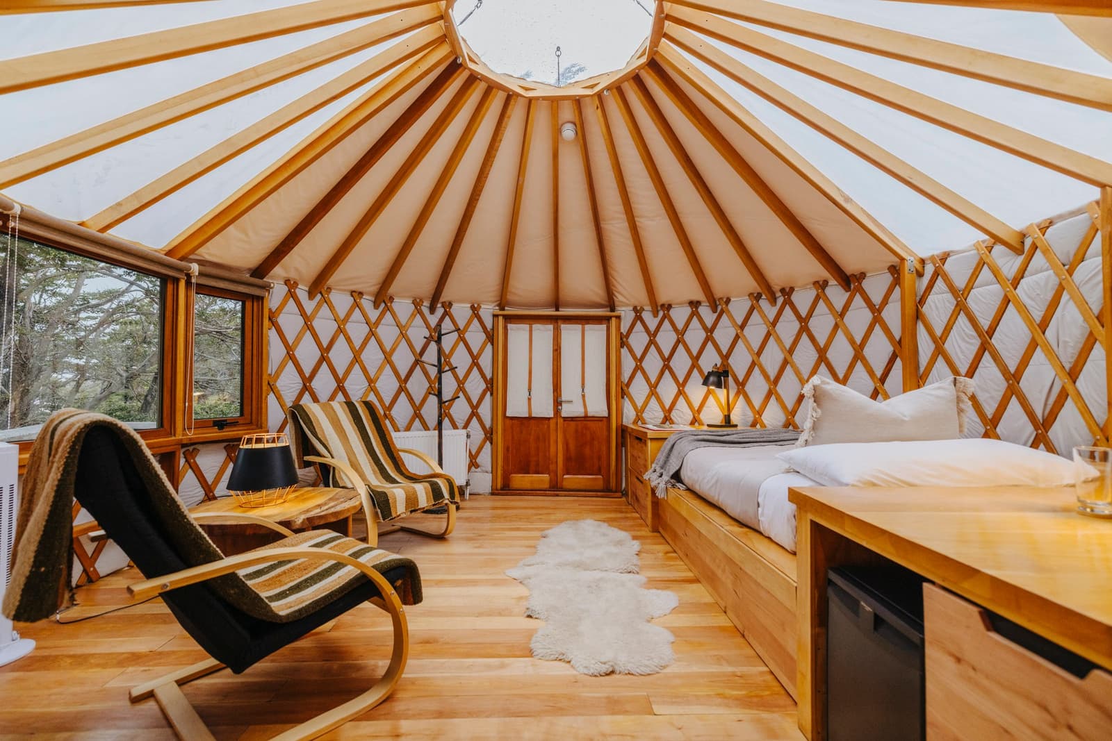Round walls, a skylight crown, and a bed that makes early mornings a genuine negotiation. Photo: Patagonia Camp Interior of a patagonia camp yurt showing the radial wooden lattice walls and canvas roof, a queen bed with white linen and a sheepskin rug on the hardwood floor, two striped rocking chairs, a bedside lamp, and a window framing green trees outside