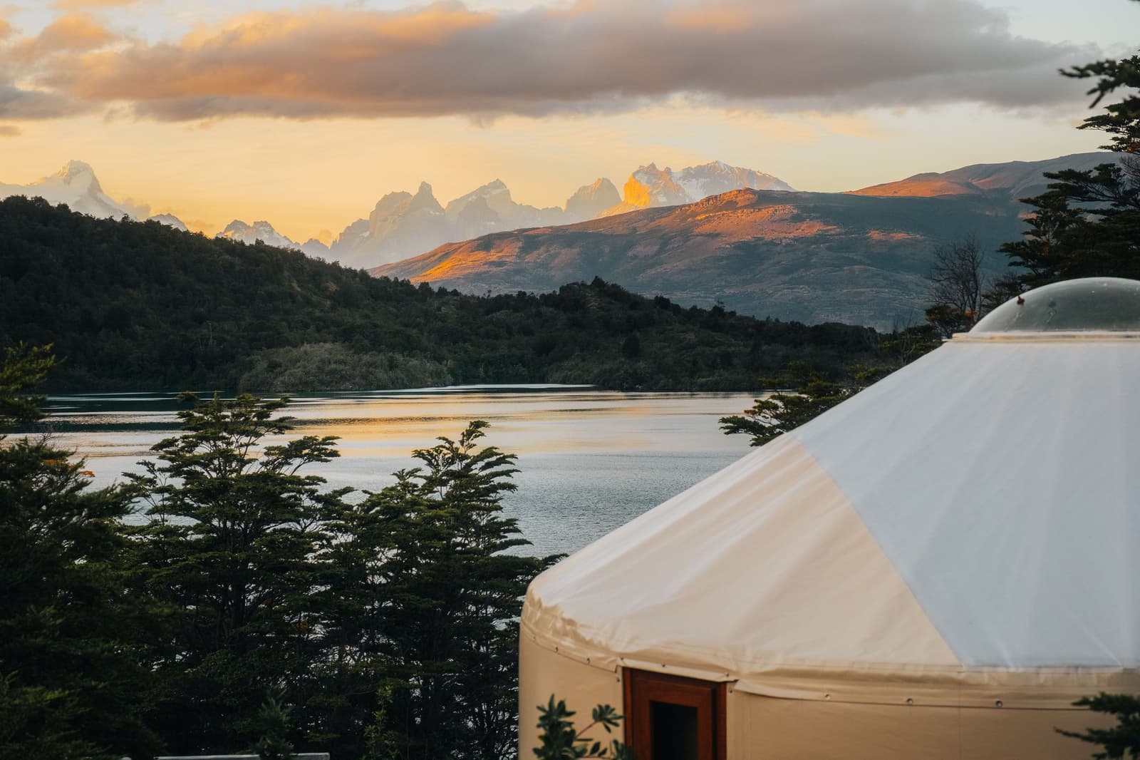Your view before dinner, no upgrade required. Photo: Patagonia Camp A white luxury yurt at Patagonia Camp in the foreground with Lake Toro and the snow-capped peaks of the Paine Massif glowing orange at sunset behind a fringe of native forest